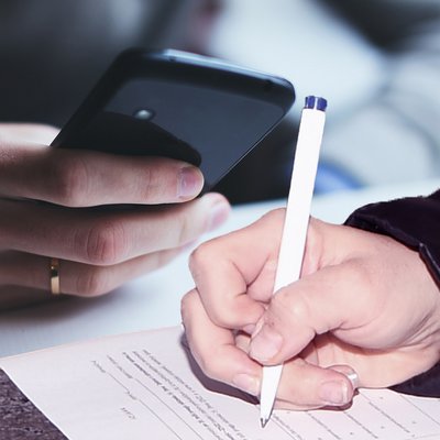 Simbolna fotografija z roko, ki drži pisalo in izpolnjuje obrazec; zraven roka druge osebe, ki drži telefon